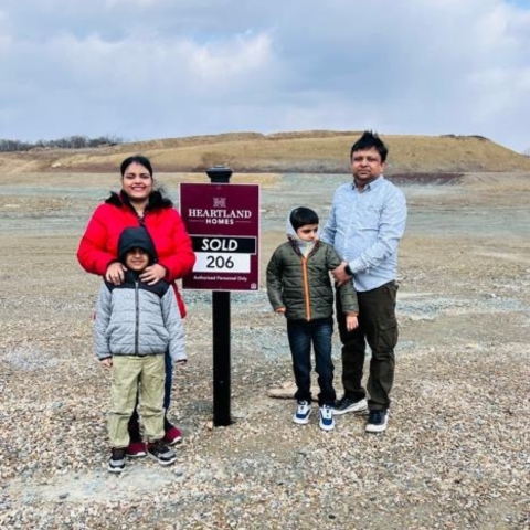 Family of four celebrating new home purchase on Heartland Homes lot, standing on gravel lot with 'SOLD' sign